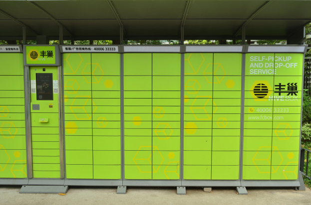 Eine grüne und gelbe Verkaufsmaschine mit Dach und Text darauf, die neben einer Straße steht und von einem Metallzaun, Pflanzen, Bäumen und einem bewölkten Himmel umgeben ist und als Selbstabhol- und -bringdienst dient.