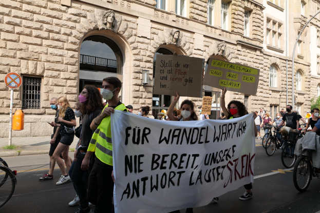 Eine Gruppe von Menschen marschiert auf einer Straße, einige halten Schilder und andere fahren Fahrröder, im Hintergrund ein Gebäude mit Fenstern, Überdachungen, Pfeilern und Skulpturen.