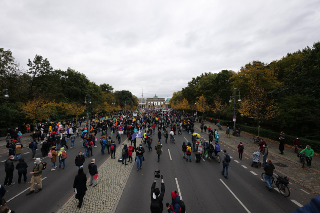 Große Menschenmenge marschiert auf einer von Bäumen gesäumten Straße in Berlin, einige halten Kameras, mit einem Gebäude und einem klaren Himmel im Hintergrund.