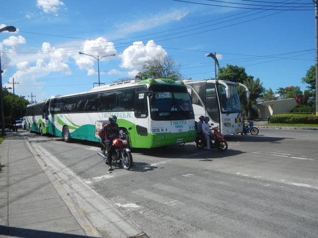 Ein grüner und weißer Shuttlebus steht am Straßenrand mit ein paar Motorradfahrern davor, ein Fußweg mit Gras und Pflanzen links daneben und Laternenpfähle, Bäume, Gebäude und einen klaren blauen Himmel im Hintergrund.