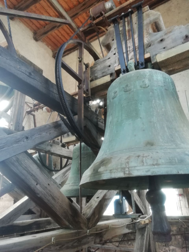 Eine große Glocke hängt an der Seite eines Gebäudes, umgeben von hölzernen Pfählen mit einer Wand im Hintergrund.