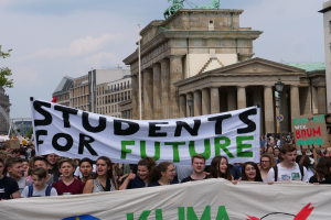 Eine Gruppe von Schülern marschiert in Berlin, hält eine bunt bemalte Plakette, auf der 'Schüler für die Zukunft' steht, mit Gebäuden, Bäumen und Himmel im Hintergrund.