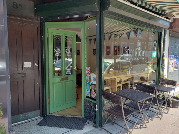 Außensitzbereich des Green Kitchen Vegan Cafe mit Tischen, Stühlen, Pflanzen, einer Fußmatte und einer Glastür mit Postern, die in einen Innenraum mit Tischen, Stühlen und einer Hängelampe führen.