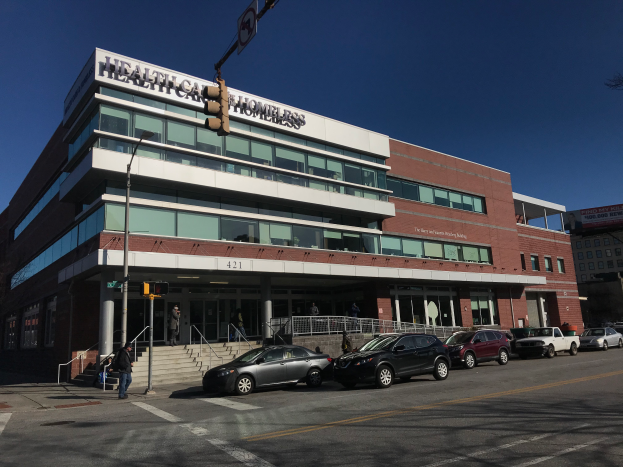 Ein großes Gebäude, das als das New Yorker Gesundheitszentrum für Obdachlose identifiziert wurde, mit Autos davor, Straßenlaternen, Lichtmasten, Verkehrsampeln, Schildern, Menschen auf dem Gehweg, Geländern, Treppen, Bäumen und einem klaren blauen Himmel.