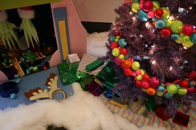 Eine Indoor-Szene mit einem geschmückten Weihnachtsbaum rechts, Geschenkboxen und Deko-Artikeln in der Mitte und einer Hütte mit Dekoration oben links, mit Watte und Wolle unten.