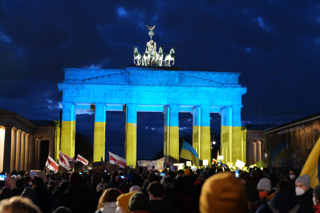 Eine Menge steht vor dem Reichstagsgebäude in Berlin, hält Fahnen und Schilder, mit einem Banner auf der rechten Seite mit protestrelevanten Text.