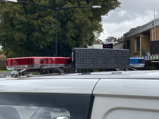 Ein Streifenwagen mit eingeschaltetem Blaulicht steht auf der Seite einer Straße, umgeben von Gebäuden, Bäumen und Masten, mit Texttafeln in der Nähe, unter einem bewölkten Himmel, wahrscheinlich nach einem tödlichen Schulschießen in Brisbane.