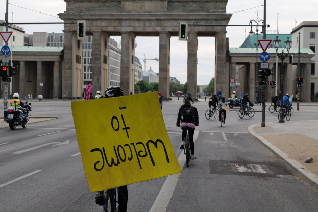 Eine Gruppe von Radfahrern mit Helmen fährt auf einer Straße am Brandenburger Tor in Berlin, Deutschland vorbei, wobei eine Person ein gelbes Schild hält, Lichtmasten, Verkehrsampeln, Gebäude, Bäume und einen klaren blauen Himmel im Hintergrund zu sehen sind.