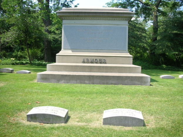 Ein Denkmal umgeben von Steinen, Gras, Bäumen mit sichtbarer Rinde, Pflanzen und einem bewölkten Himmel.