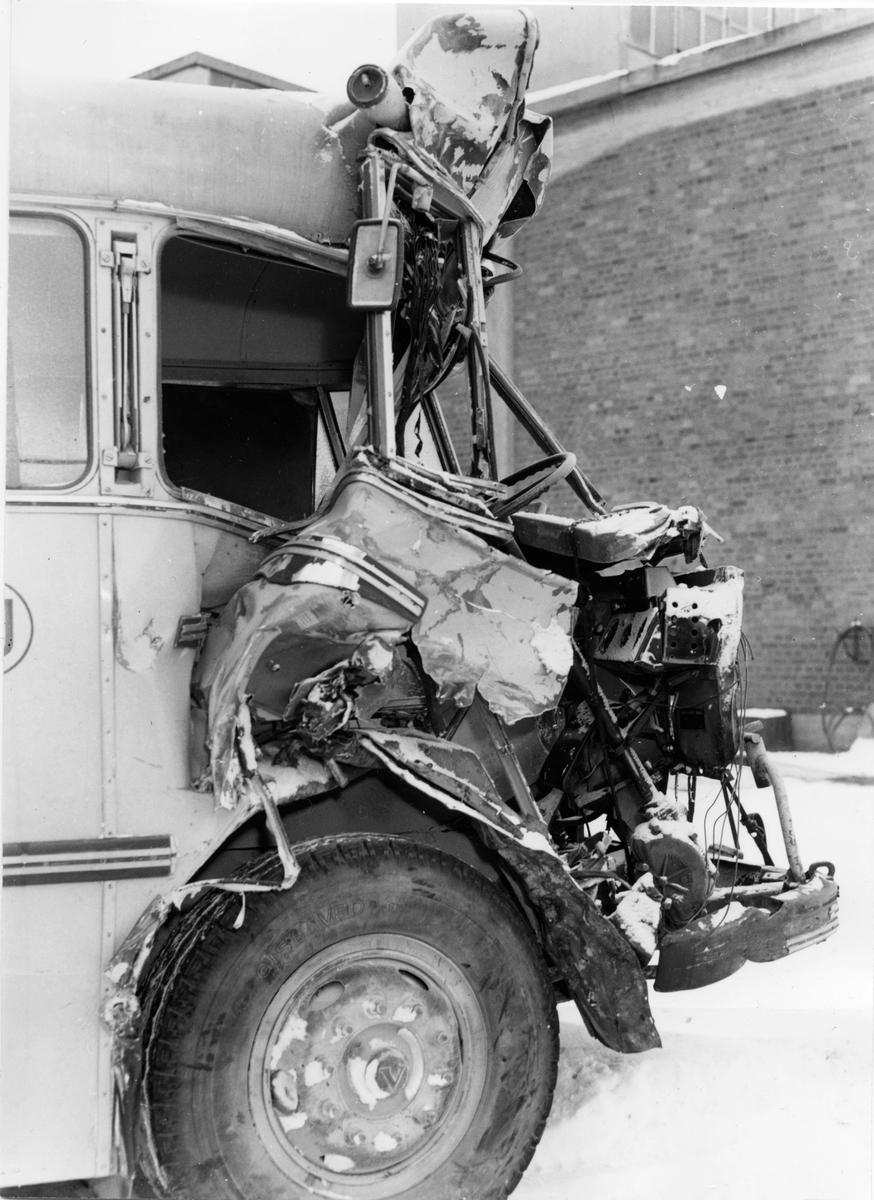 Ein Schwarz-Weiß-Foto eines verunglückten Busses mit schwerer Frontschaden, der auf der Seite einer Straße mit einer Wand im Hintergrund geparkt ist.