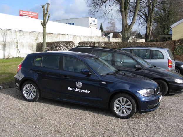 Mehrere Autos stehen auf einem Weg umgeben von grünem Gras, Sträuchern und Bäumen. Im Hintergrund gibt es Gebäude, ein Schild mit Text und ein Logo.