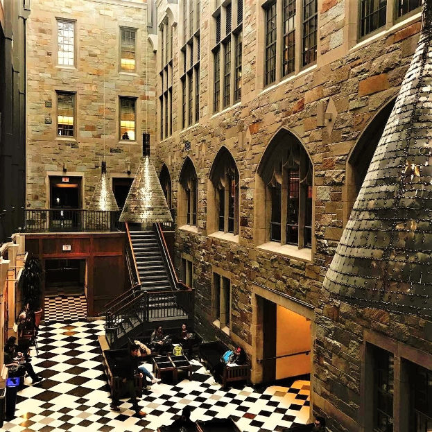 Großer Raum mit schwarz-weißem Schachbrettboden, zentraler Treppe mit Geländern, Fenstern, Deckenbeleuchtung und mehreren Personen an Tischen und Stühlen sitzend, ähnlich einer Harry-Potter-Studio-Tour.