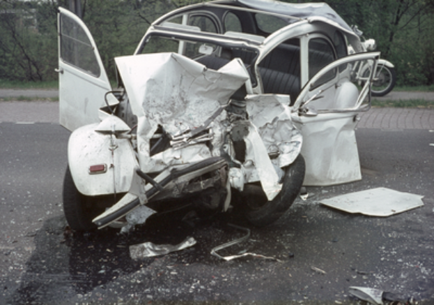 Ein stark vorne verbeultes Auto steht am Straßenrand, mit Bäumen, einem Gebäude und einem anderen Fahrzeug im Hintergrund.
