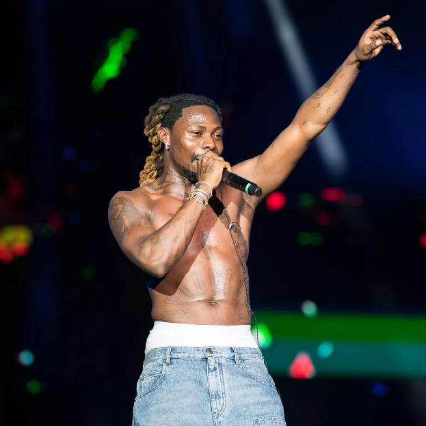 Travis Scott performs auf der Bühne des Coachella Valley Music and Arts Festival, hält ein Mikrofon und ist von hellen Lichtern umgeben mit einem unscharfen, träumerischen Hintergrund.