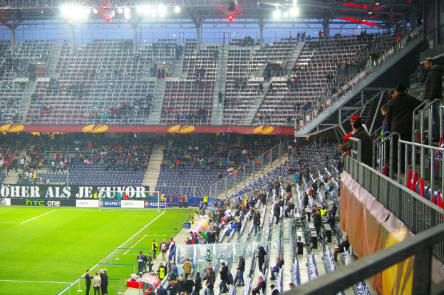 Ein großes Stadion voller Zuschauer bei einem Fußballspiel, mit einem Geländer, einer Fahne auf der rechten Seite und einem leeren Abschnitt im Hintergrund mit Deckenbeleuchtung.