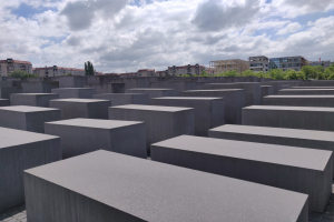 Luftaufnahme des Holocaust-Mahnmals für die ermordeten Juden Europas in Berlin, das ein Gittermuster aus Betonsteinen zeigt, mit Bäumen, Gebäuden und Wolken im Hintergrund.