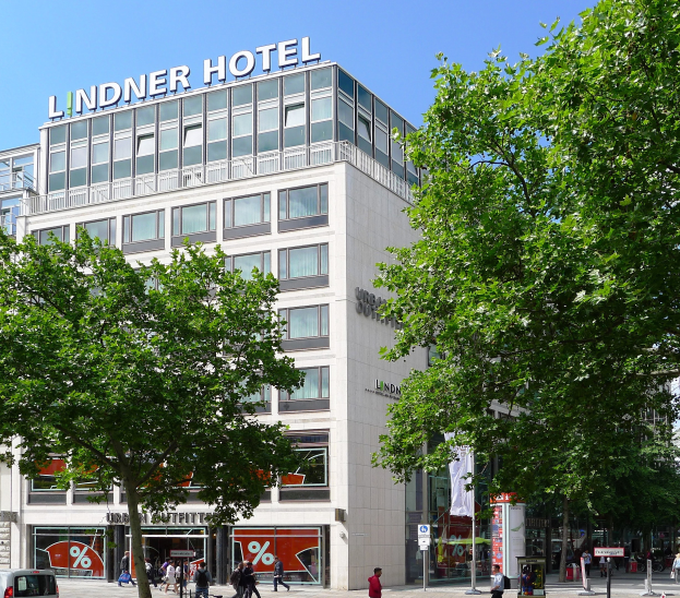 Großes modernes Hotelgebäude mit Glasfenstern und Beschriftung auf der Spitze, umgeben von Bäumen und einer belebten Straße mit Fußgängern und Fahrzeugen unter einem sichtbaren Himmel.