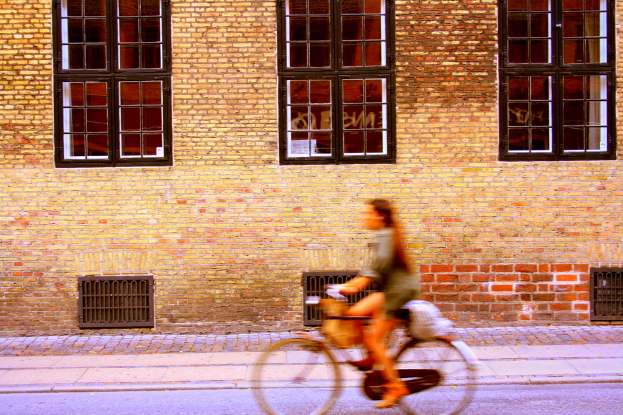 Eine Person fährt mit dem Fahrrad auf einer Straße neben einer Wand mit Fenstern und Metalltüren.