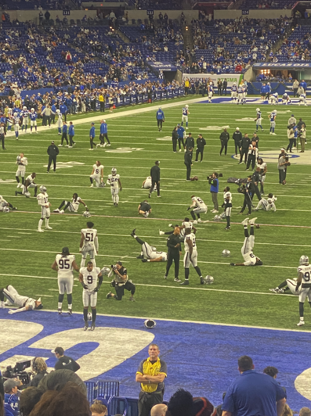 Ein Fußballspiel wird in einem Stadion gespielt, während Menschen auf dem Feld stehen und einige auf Stühlen im Hintergrund sitzen, während eine Person eine Kamera hält, was auf eine Live-Übertragung hinweist.