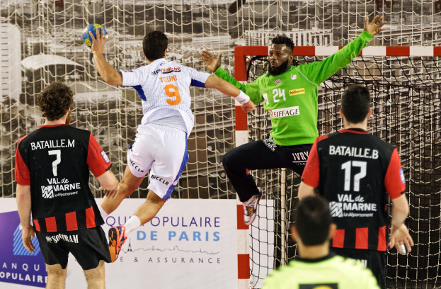 Eine Gruppe von Männern, die Handball spielen, mit einem Spieler, der springt und den Ball hält, vor einem Torpfosten und einer Fahne mit der Aufschrift 'Ligue 1 - Bordeaux vs Marseille.'