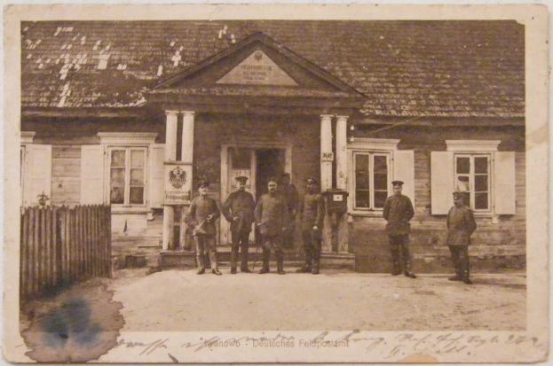 Ein Schwarz-Weiß-Foto von einer Gruppe von Männern, die vor einem Gebäude mit Fenstern, Säulen und einem Dach stehen, mit einem Zaun auf der linken Seite und einem Text am unteren Rand, der 'Deutsche Post in Berlin' lautet.