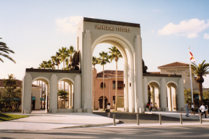 Eingang zu Universal Studios Hollywood mit einem Bogen, einer Statue, Menschen, Bäumen, Gebäuden, einem Fahnenmast und einem bewölkten Himmel.