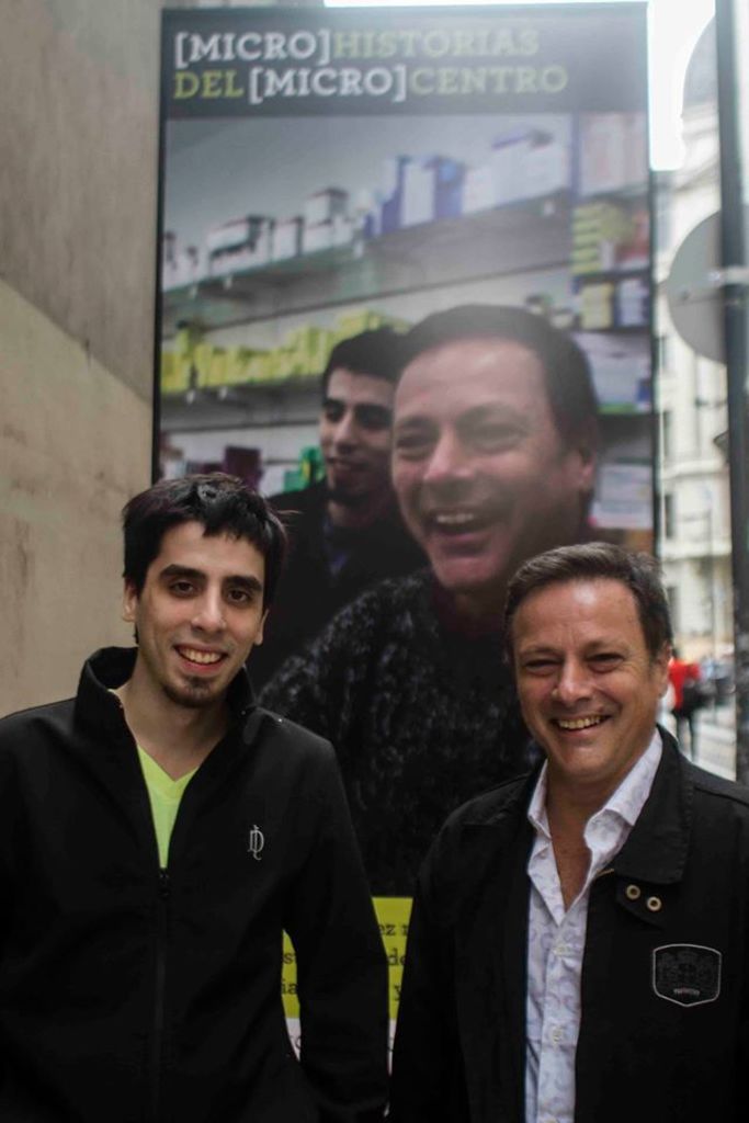 Zwei Personen posieren vor einem Plakat mit ihren Gesichtern, auf dem 'micro historias del microcentro' steht; die Person rechts trägt ein weißes Hemd und einen schwarzen Anzug, während die Person links einen schwarzen Pullover und ein grünes T-Shirt trägt.