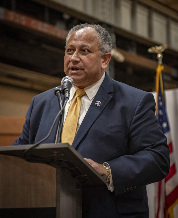 Ein Mann in Anzug und Krawatte spricht an einem Podium mit Mikrofon, eine Flagge rechts und eine Wand im Hintergrund, wahrscheinlich zur Ankündigung eines neuen Bürgermeisters von New York City.