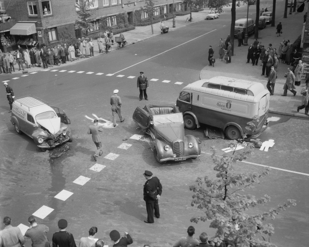 Schwarz-weiß-Bild von einem Autounfall an einer Straßenkreuzung mit einem Lieferwagen, anderen Fahrzeugen und einer Gruppe von Menschen, mit Gebäuden, Bäumen, Pfählen und Texttafeln im Hintergrund.