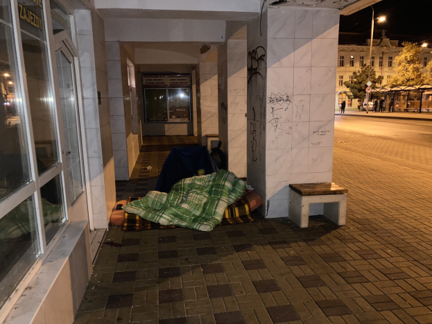Eine obdachlose Person, die auf einem Bürgersteig bei Nacht schläft, mit einer Decke und einem Hocker in der Nähe, vor einem Gebäude mit einer Schautafel, einem Straßenpfahl, Bäumen, Fahrzeugen, Passanten und anderen Gebäuden unter einem bewölkten Himmel.