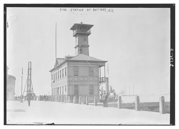 Ein Schwarz-Weiß-Foto einer Feuerwache in Battery, New York, mit einem Gebäude, Menschen, Pfählen, einem Zaun, Booten auf dem Wasser und einer bewölkten Himmel.