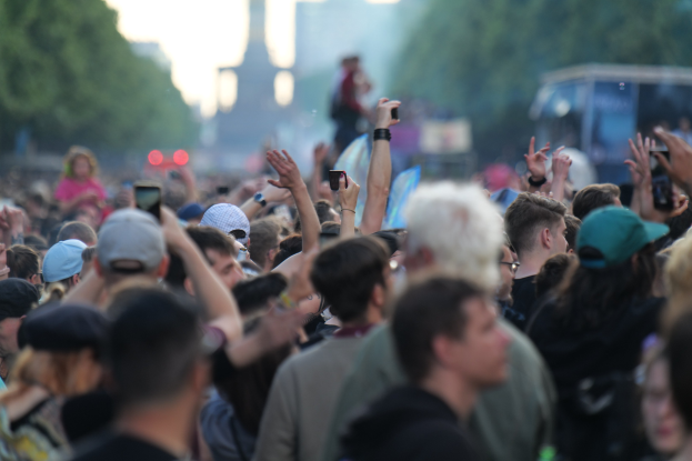 Menschenmenge auf einem Musikfestival mit erhobenen Händen, einige tragen Käpchen und halten Telefone, vor einem unscharfen Hintergrund von Bäumen, Gebäuden, Lichtern und einem Fahrzeug.