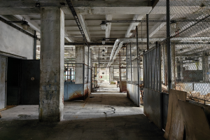 Leerer Raum mit großen Fenstern, Säulen, einer Tür, Deckenleuchten, Metallstangen, einem Maschendrahtzaun, verstreuten Gegenständen auf dem Boden und einer Wand, die eine verlassene Fabrik mit Anzeichen von Vernachlässigung zeigt.