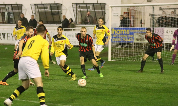 Ein Sportplatz mit mehreren Männern, die Fußball spielen, ein Netz auf der rechten Seite, eine Tafel mit Text im Hintergrund und eine Gruppe von Zuschauern, die hinter der Tafel stehen, mit einer Wand und Fenstern im Hintergrund.