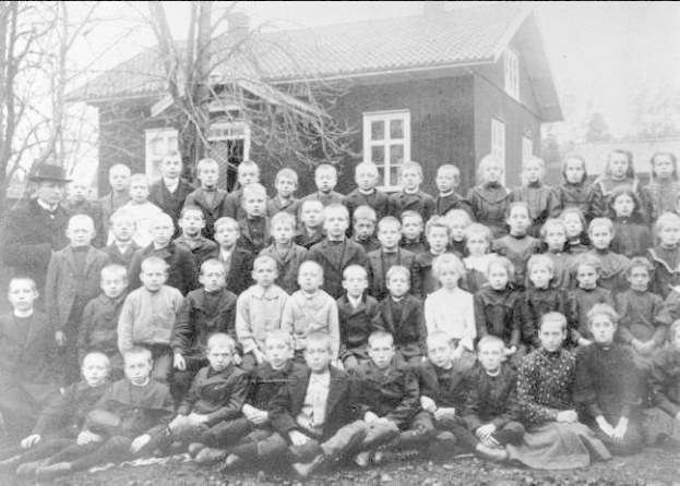 Schwarz-weiß-Foto einer Gruppe von Kindern, die vor einem Haus mit Fenstern posieren, umgeben von Bäumen und einem klaren Himmel, mit dem Text "1930er Schulkinder" unten.