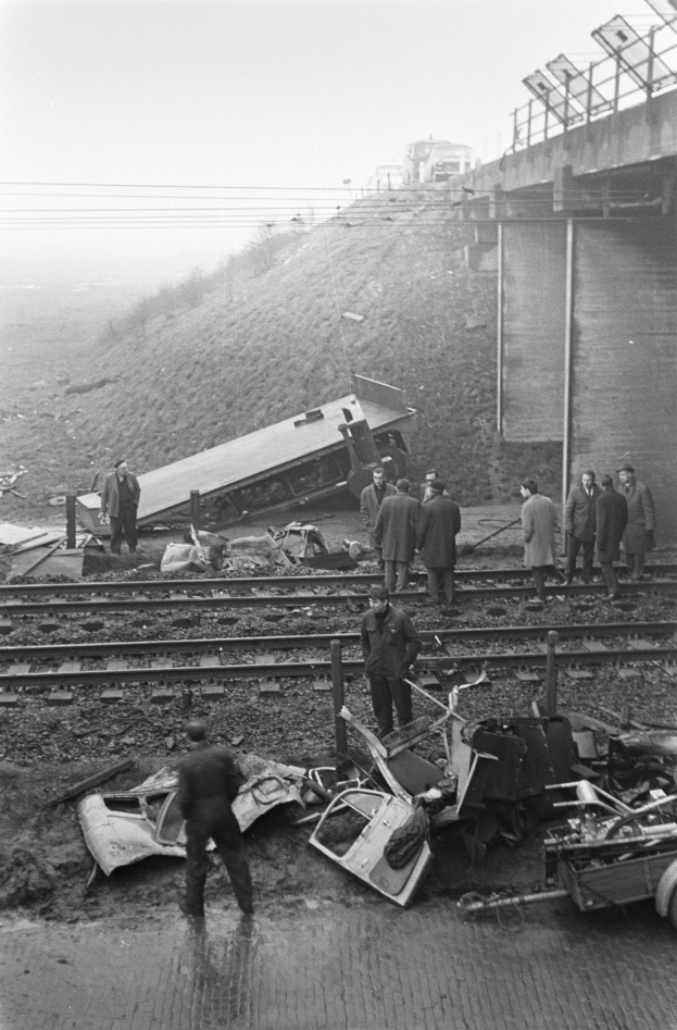Schwarzes und weißes Bild einer Zugentgleisung mit Trümmern auf den Gleisen, einer Gruppe von Menschen in der Nähe, einem Fahrzeug im Vordergrund und einer Brücke, Masten, Drähte und Himmel im Hintergrund.