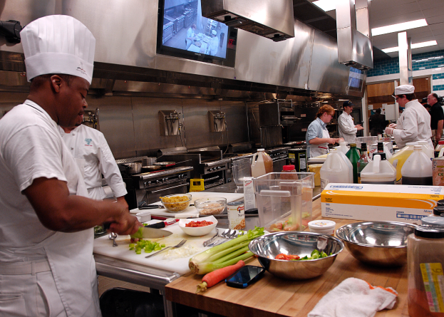 Eine Gruppe von Köchen in einer Küche, die auf einem Schneidbrett Lebensmittel zubereiten, mit einem Koch im Vordergrund in einer weißen Schürze und Mütze, der ein Messer hält und von Utensilien, Schüsseln, Gemüse, einem Handy, Flaschen und anderen Gegenständen umgeben ist, mit einem Fernseher und Deckenlampen im Hintergrund.