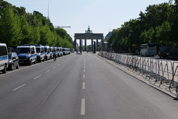 Eine Reihe von Polizeiwagen, die auf einer Straße vor dem Reichstagsgebäude in Berlin, Deutschland, geparkt sind, mit Menschen auf Fahrrädern und Stehenden, Barrieren, Bäumen und einem Bogen mit Statuen im Hintergrund.