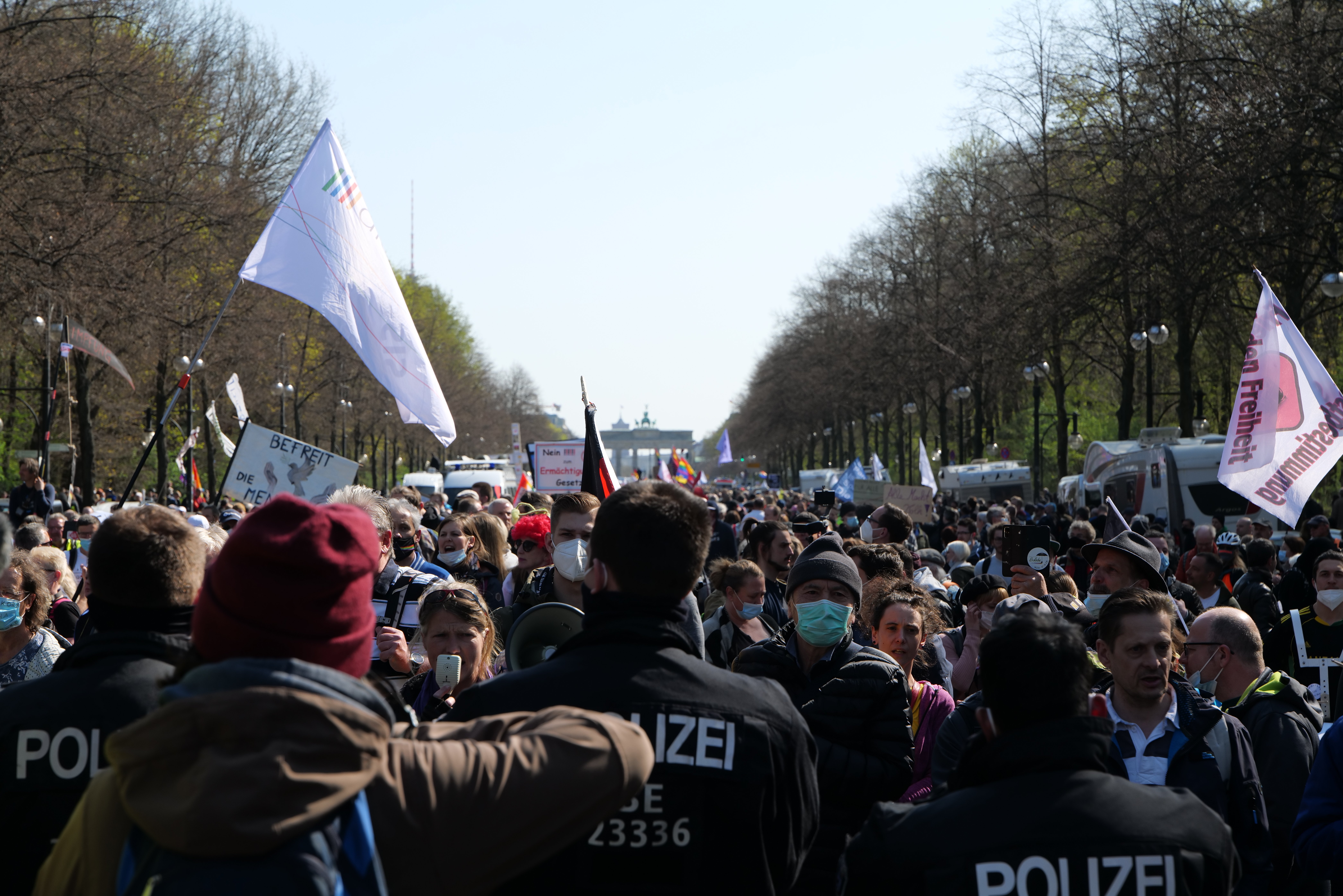 Eine große Gruppe von Menschen steht vor einer Menge von Polizisten, einige tragen Mützen und Masken, während sie auf einer Demonstration in Berlin, Deutschland, mit Schildern, Fahnen, Laternenpfählen, Bäumen, Fahrzeugen, einem Gebäude und dem Himmel im Hintergrund demonstrieren.