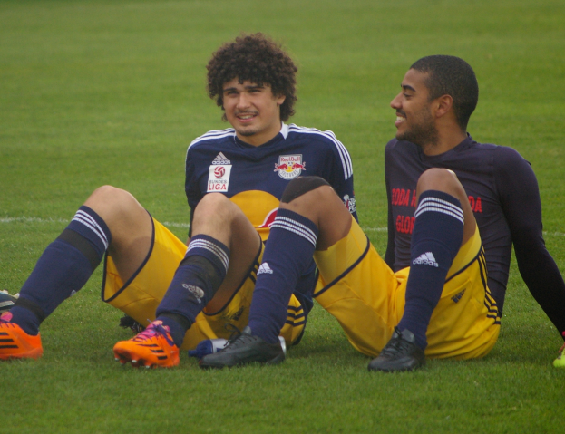 Zwei Fußballspieler sitzen auf dem Boden mit übereinandergeschlagenen Beinen, tragen Sportbekleidung und lächeln sich an.