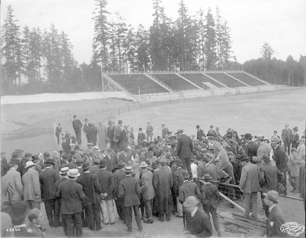 Ein Schwarz-Weiß-Bild einer Menge, die sich um ein Fußballfeld versammelt hat, während der Eröffnungszeremonie der Olympischen Spiele 1904, mit Bäumen und einem klaren Himmel im Hintergrund und Text mit einem Logo unten.