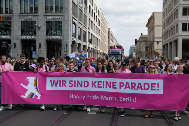 Eine Gruppe von Menschen marschiert auf einer Straße in Berlin mit einem pinken Banner, auf dem "Happy Pride March" steht, vorbei an Gebäuden, Laternenpfählen und Verkehrszeichen unter einem bewölkten Himmel.