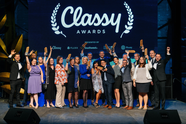 Eine Gruppe von Menschen auf einer Bühne, die Auszeichnungen halten, mit Lautsprechern darunter und einem Bildschirm im Hintergrund, der "Classy Awards 2019" anzeigt.