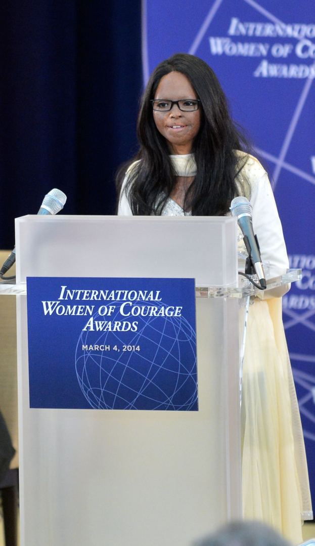 Eine Frau in einem weißen Kleid und Brille steht an einem Podium mit zwei Mikrofonen und einer Tafel, während sie eine Menge bei den "International Women of Courage Awards 2014" anspricht.