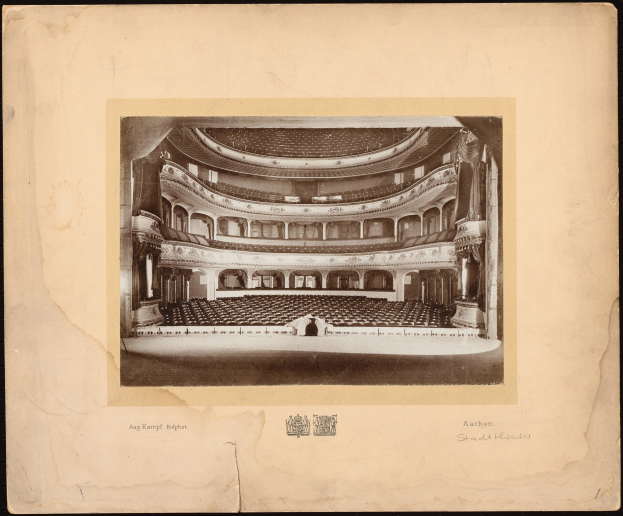 Ein altes Schwarz-Weiß-Foto des Auditoriums der Royal Albert Hall in London, das die Bühne mit Reihen von Stühlen, Säulen, Geländern und Text und einem Logo unten zeigt, mit einem dunklen Hintergrund.