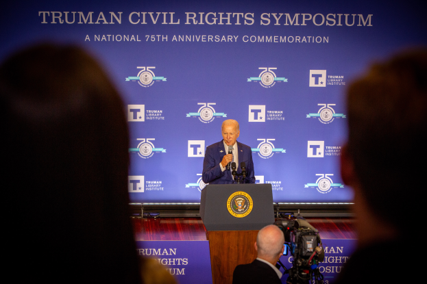 Ein Mann im Anzug und mit Krawatte steht an einem Rednerpult und hält ein Mikrofon in der Hand, hinter ihm eine Fahne mit der Aufschrift "Trumpan Civil Rights Symposium", während rechts von ihm jemand eine Videokamera bedient.