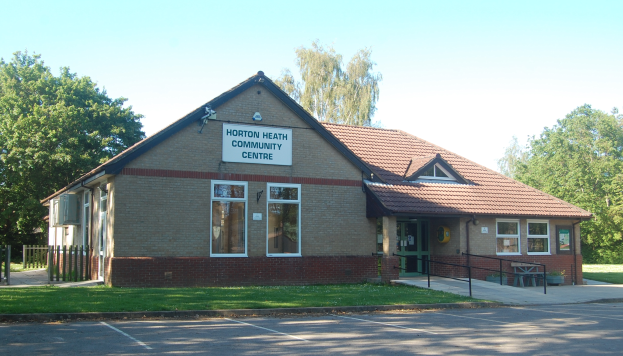 Ein Haus mit einem Dach, Fenstern, Türen, einem Schild mit der Aufschrift "Horton Heath Community Centre", einem Metallzaun, einer Bank, Topfpflanzen, Gras, einem Weg, einer Baumgruppe und einem bewölkten Himmel.