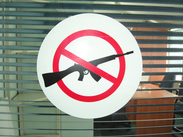 Ein Schild mit der Aufschrift 'Keine Waffen' auf einem Bürofenster mit einer Tafel, die ein Gewehrbild zeigt, Stühle, einen Tisch und eine Wand im Hintergrund.