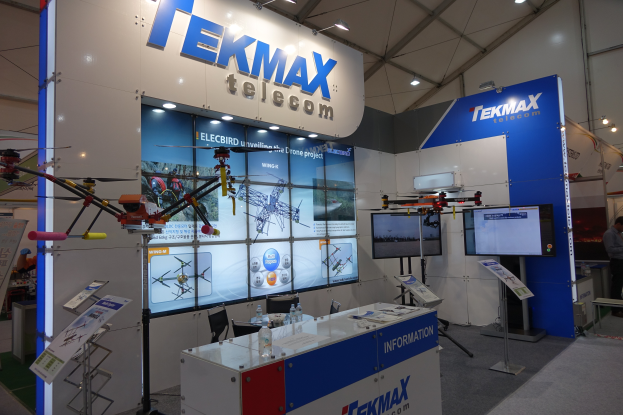 Messestand für Tekmax Telecom mit großem Wanddisplay, Tisch mit Produkten, zwei Fernsehern, Infotafeln, Deckenbeleuchtung und einigen Personen.