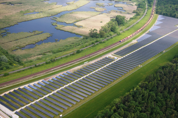 Luftaufnahme einer Solaränlage mit Panelen in einem Feld, umgeben von Bäumen, Gras, Wasser und einem vorbeifahrenden Zug.